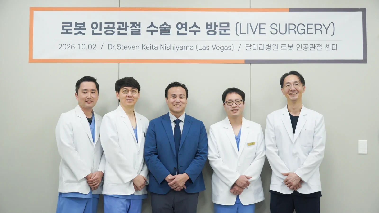 로봇 인공관절 수술 연수 방문(LIVE SURGERY) 현수막 아래에서 파란 정장 차림의 외국인 의사와 흰 가운의 달려라병원 의료진 4명이 함께 선 단체 사진