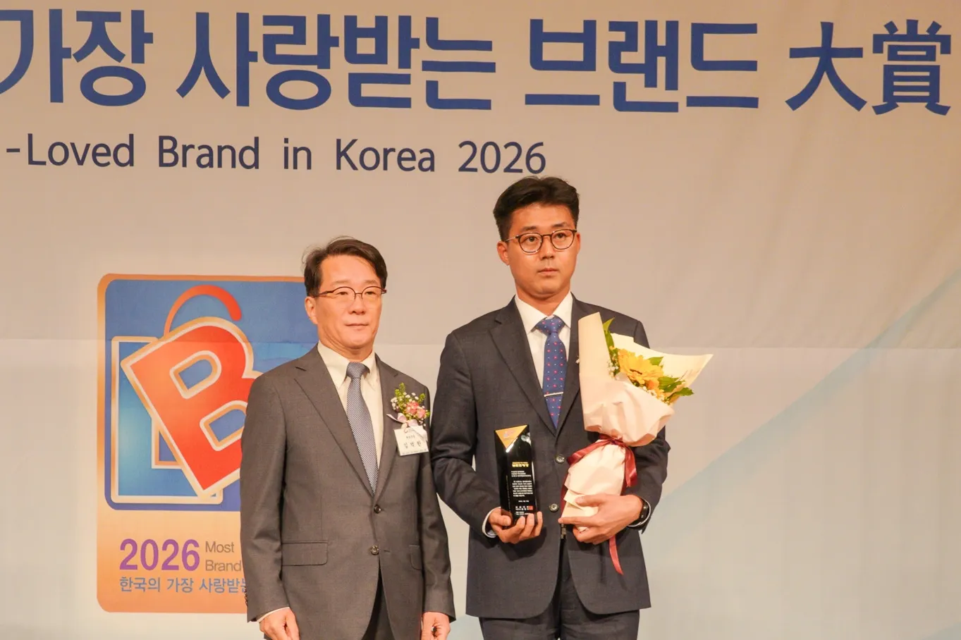 2026 한국의 가장 사랑받는 브랜드 大賞 시상식에서 척추관절병원 부문 대상을 수상한 달려라병원 관계자가 무대에 서서 트로피와 꽃다발을 들고 시상자와 나란히 있는 사진. 뒷배너에 "Most-Loved Brand in Korea 2026" 문구와 2026 로고가 보인다