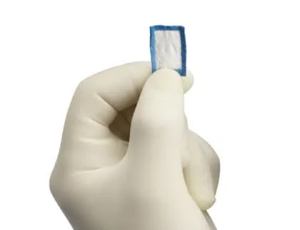 수술용 장갑을 낀 손이 리제네텐 패치를 손가락으로 집어 들고 있는 사진. 패치는 손가락 한 마디 남짓한 크기의 흰색 사각형이며 가장자리가 파란 실로 둘러져 있다